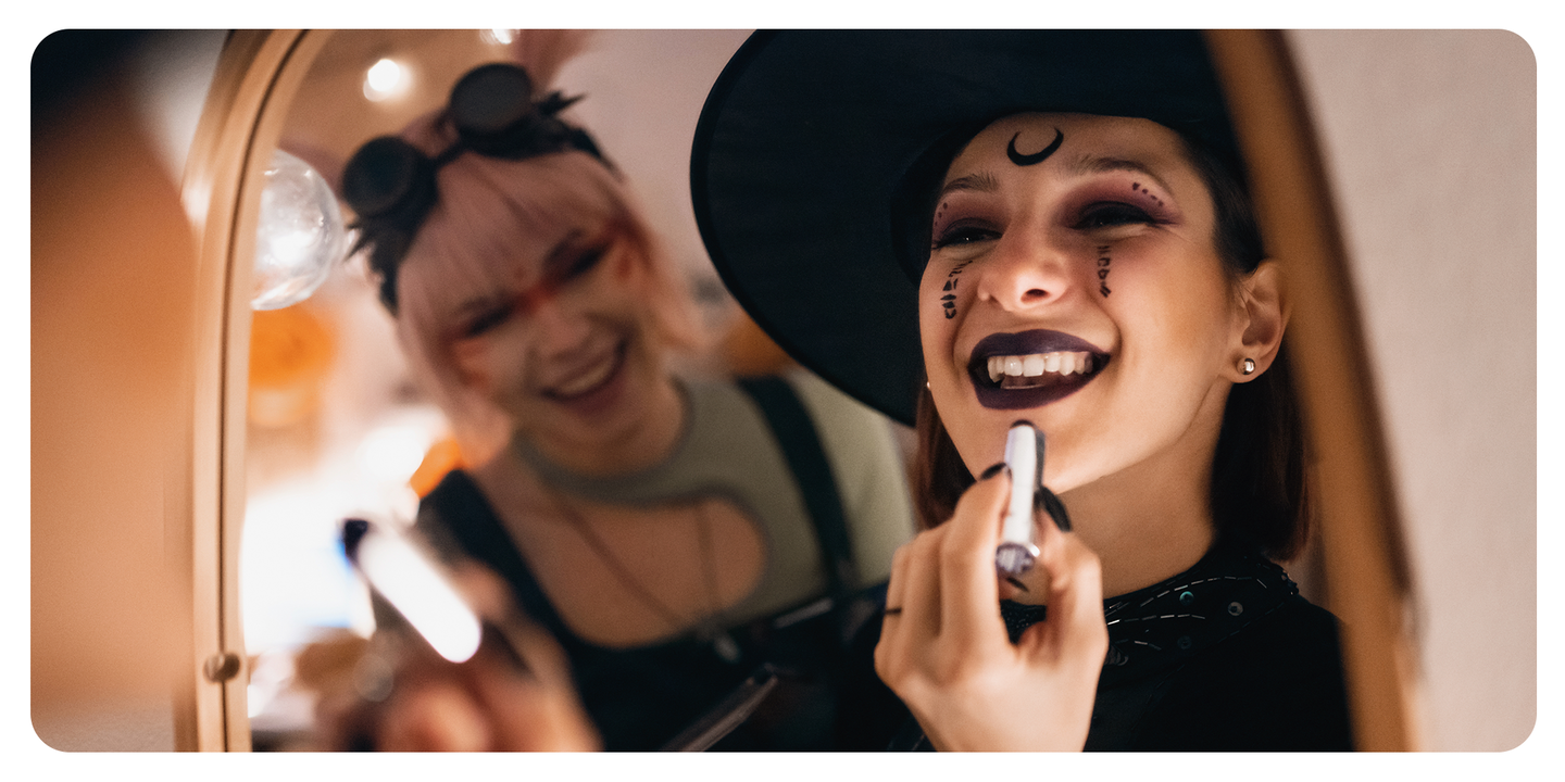 Two women in witch costumes applying Halloween makeup and dark lipstick in a mirror